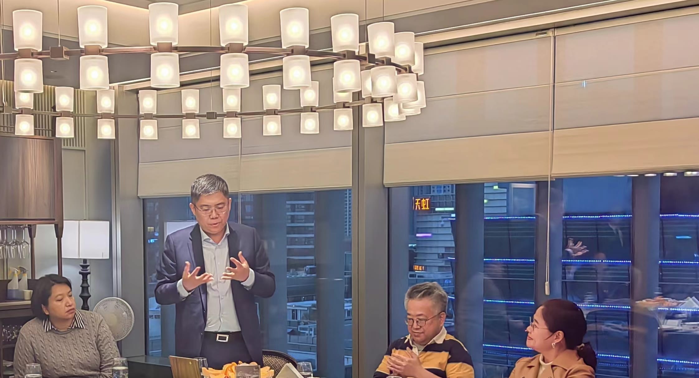 Man in suit speaking at table, overhead chandelier, city view visible through window.